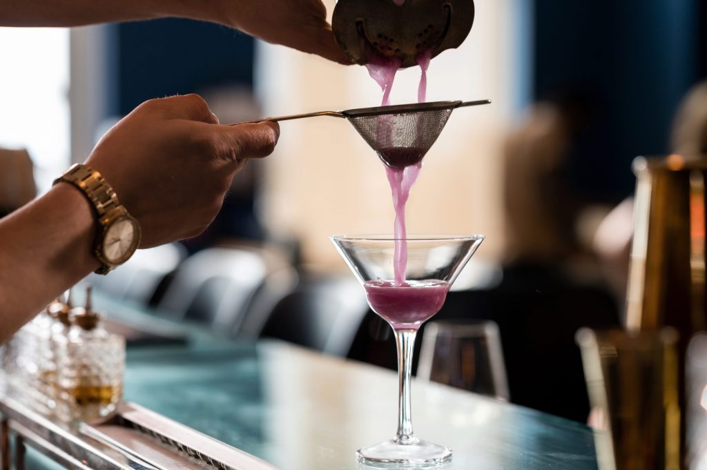 Bartender straining a purple cocktail into a martini glass at Gather