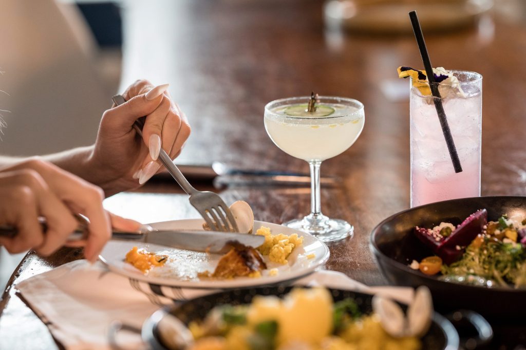 Guest cutting into a dish beside a coupe cocktail and a pink highball at Gather