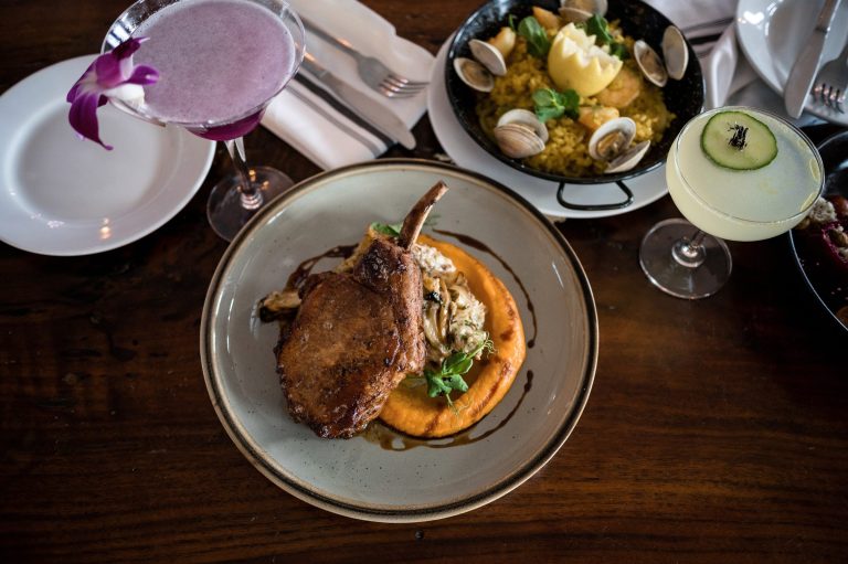 Overhead view of a bone-in chop, seafood paella and two cocktails on a Gather table