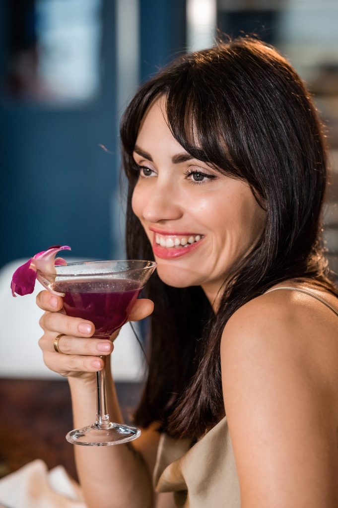 Smiling guest holding a purple cocktail garnished with an orchid at Gather