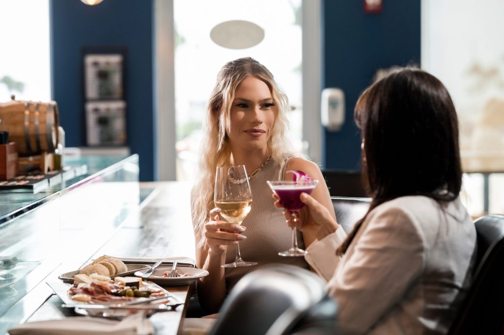 Two guests raising a glass of white wine and a purple cocktail at the Gather bar beside a charcuterie board