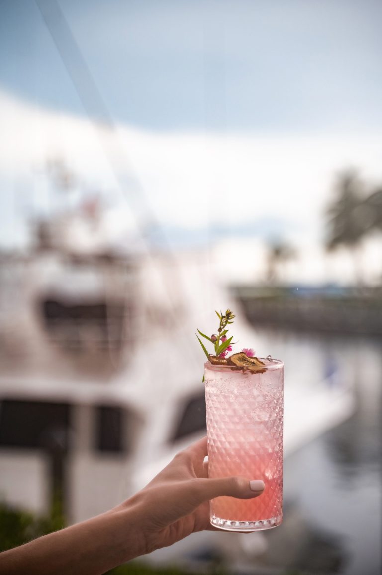 Guest holding a pink cocktail with a floral garnish in front of a yacht at the marina.