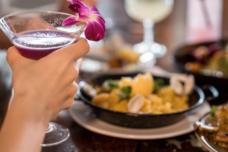 Purple cocktail with an orchid garnish raised in front of a pan of seafood paella